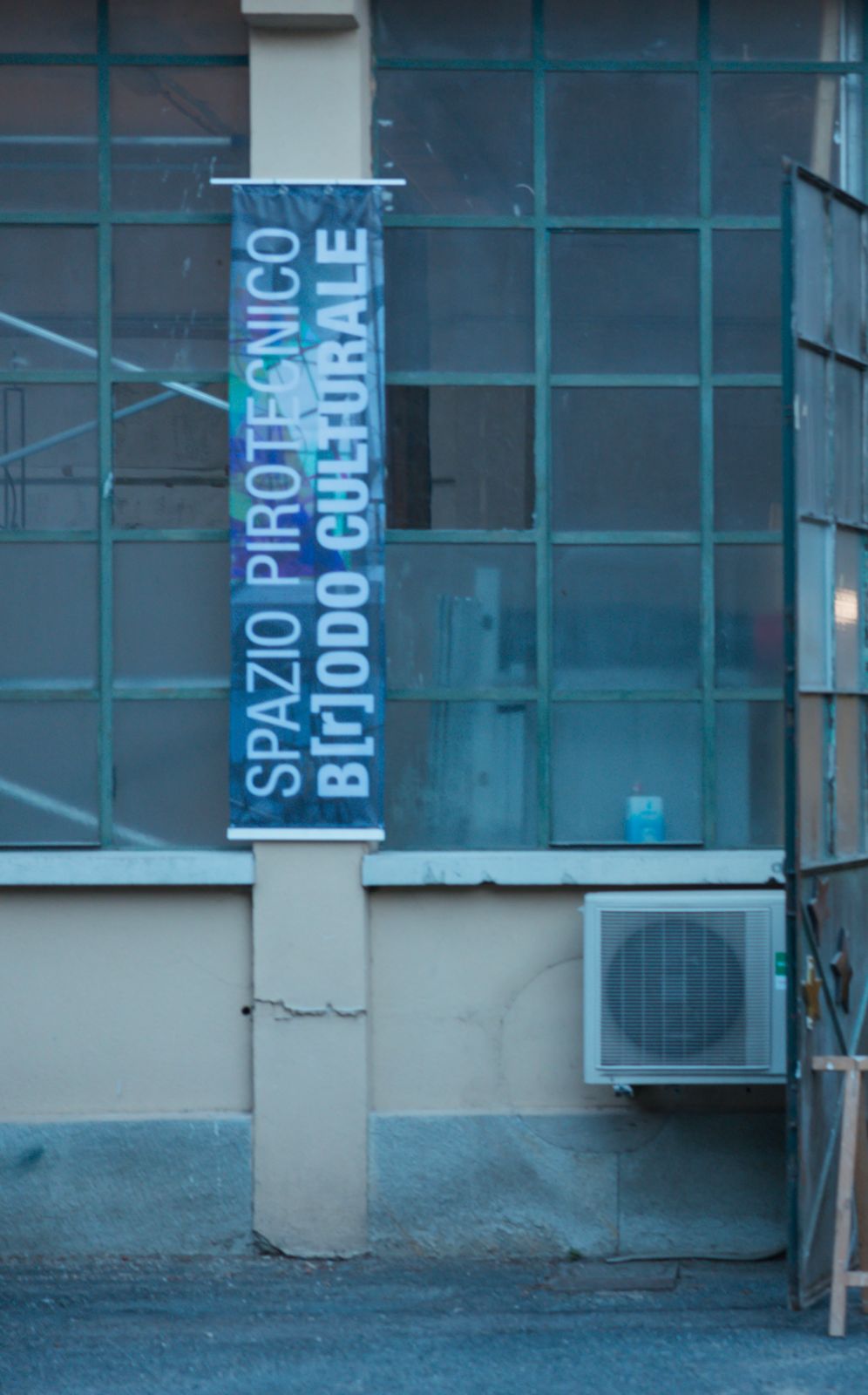 Facciata con vetrate industriali e lo striscione verticale Spazio Pirotecnico – B[r]odo Culturale
